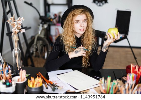 Portrait of romantic blonde long hair curly female artist in black hat sitting behind the table with color pencil and green apple