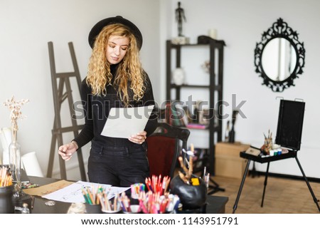 Romantic blonde long hair curly female artist in black hat stay near the table with color pencil holding sketch in her studo