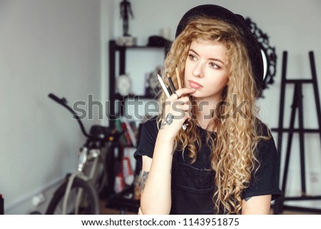 Portrait of contemplative tattoed curly painter girl in a black hat wears eyeglasses hold pencils