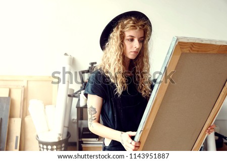 Attracive long hair artist girl in a nice black hat and holds canvas in light sunny studio