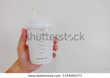 Close up: Paper coffee cup in left hand with white wall.