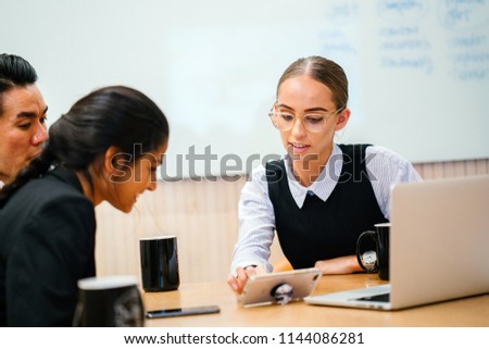 A young Caucasian professional woman has a business discussion with her diverse team in a meeting room. They are talking animatedly. There is a whiteboard in the background with a laptop on the table.