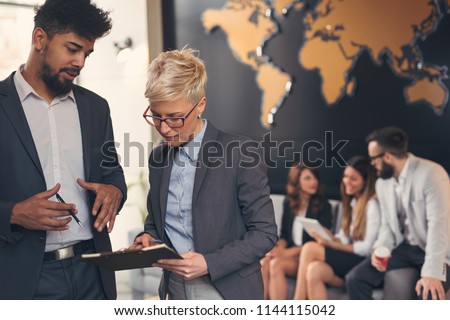 Businessman and businesswoman reviewing contract; business team working in the background. Focus on the man standing Royalty-Free Stock Photo #1144115042