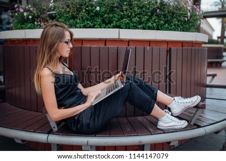 Relaxed young girl is studying in the park, lying on the wooden bench browsing on her laptop, in casual comfortable outfit outdoors in the street