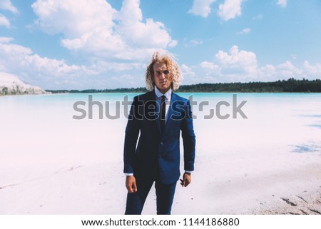 Portrait of blond curly man dressed blue business suit. Clear water and sky on the background. Summer time. Travel topic.