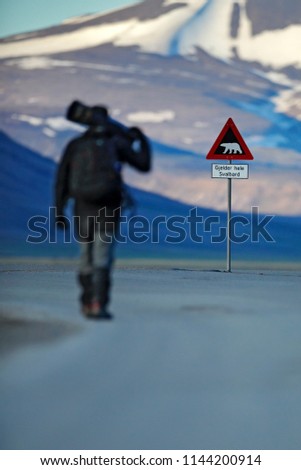 Photographer with big lens and road traffic sign with Polar bear. “Gjelder Hele Svalbard” means “Over All of Svalbard (watch out for polar bears)”. Man on the road with snowy mountain, Svalbard.