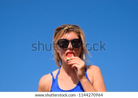 A woman is holding a piece of watermelon near her mouth against the blue sky. A ripe watermelon in the hands of a blonde.