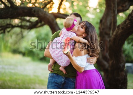 Mom and Dad hug and kiss their little daughter. Against the background of the old tree and the sunset