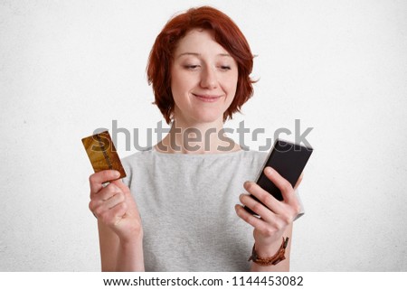 Happy delighted female with short hair, holds smart phone and plastic card, checks her bank account, being satisfied to recieve salary, uses online application, isolated over white concrete wall