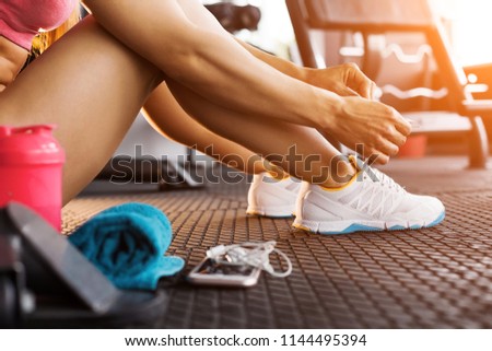 close up photo of woman tying shoelaces of white trainers siting on a mat surrounded by smart phone, earphones, towel and a shaker in the sun lightened gym Royalty-Free Stock Photo #1144495394