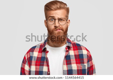 Photo of bewildered puzzled man with thick ginger beard and mustache, raises eyebrows, looks doubtfully at camera, wears fashionable clothes, isolated over white background. Facial expressions