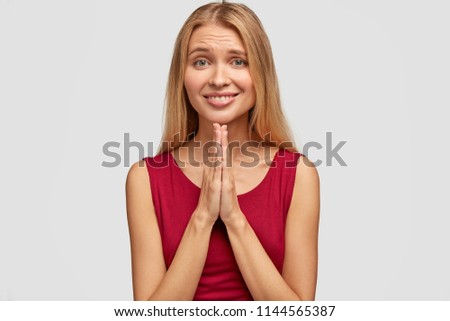 Studio shot of pleased blonde female with light hair, asks for help, keeps hands in praying gesture, has appealing look and charming smile, wears casual red t shirt, stands against white wall