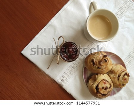 Jar of sweet strawberry jam with cup of coffee and delicious cinnamon rolls (cinnabons, cinnamon powder buns) on the plate on the wooden table with white cotton tablecloth.