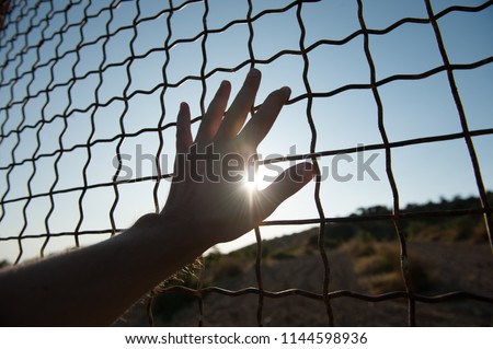 male human hand touching fence cage in jail and illegal immigration on sunset Royalty-Free Stock Photo #1144598936