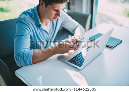 Young man freelancer checking notification on smartphone while working remotely at netbook using wireless free internet.Hipster guy chatting online in social networks on telephone sitting at laptop