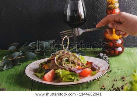 Salad with beef steak, vegetables and pesto sauce in rustic plate on green and black wooden background with ingredients and glass of wine
