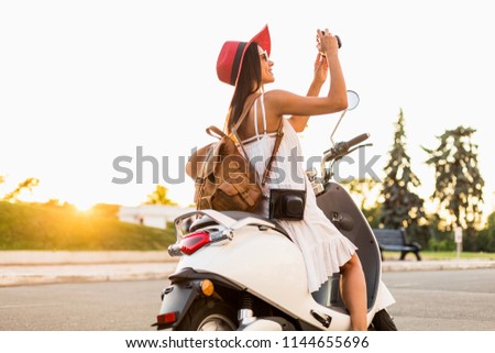 attractive woman riding on motorbike in street, summer vacation style, traveling, smiling, having fun, stylish outfit, adventures, taking pictures on vintage photo camera, wearing leather backpack