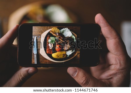 Food or technology concept. Man hands taking food photo by mobile phone. Close up of male hands holding modern smartphone, tasty dinner on the background. Toned image. Selective focus.