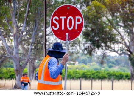 Unrecognizable man with safety vest and hat is holding a stop sign with his hands on the street for construction work. There are many workers like this in Australia for construction works.