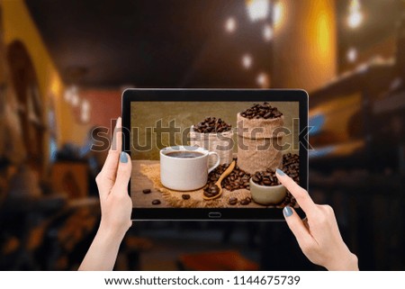 Hand using mock up computer tablet, with picture hot cup of coffee with beans on burlap sack on coffee shop cafe background, and copy space