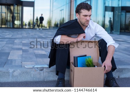Fired business man sitting frustrated and upset on the street near office building with box of his belongings. He lost work Royalty-Free Stock Photo #1144746614