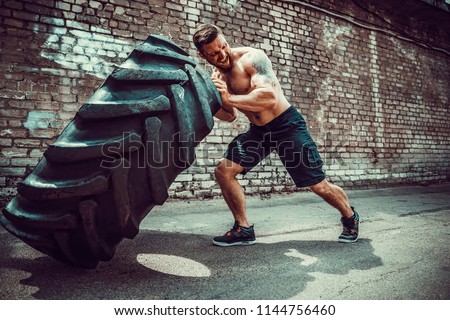 Muscular bearded tattooed fitness shirtless man moving large tire in street gym. Concept lifting, workout training. Royalty-Free Stock Photo #1144756460