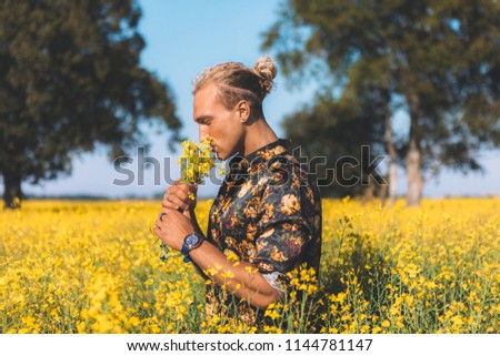 Portrait of curly blond man. Standing at the field and holding a flowers. Trees around, romantic, delicate photo. Summer time.
