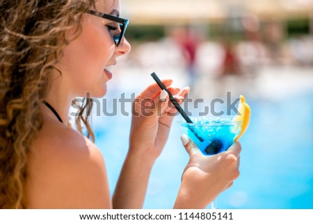 close up face profile portrait of young beautiful caucasian woman with long brown hair wearing sunglasses drinking blue cocktail though a straw over outdoor poolside area blurred bckground. Royalty-Free Stock Photo #1144805141