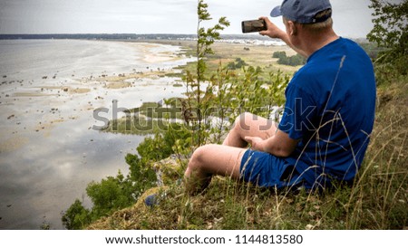 Young man with a smartphone and baseball cap taking a photo of sea and hills while trekking at sunset