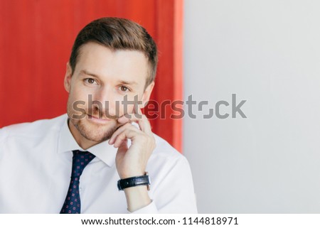 Self assured unshaven male office worker in formal white shirt with tie, holds hand on cheek, thinks about starting new stage in life, poses against red and white background. Business concept