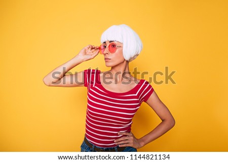 Beautiful young hipster woman wearing blonde wig and pink sunglasses posing against orange background