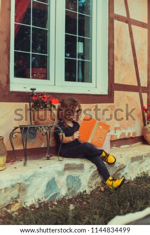 A little pretty blonde girl in glasses reads and yellow boots reads an old book sitting on the porch under the house in the summer
