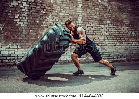 Muscular bearded tattooed fitness man moving large tire in street gym. Concept lifting, workout training. Royalty-Free Stock Photo #1144846838