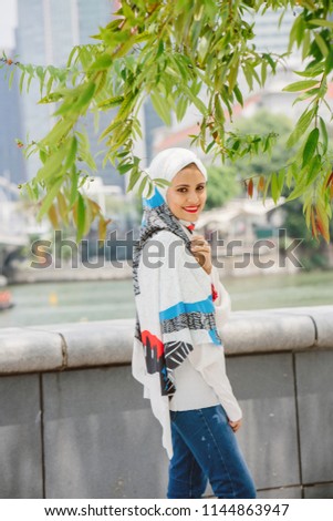 A young Muslim woman in hijab (tudung, headscarf) stands in the shade on a sunny day against the city in the background. Her head scarf is colorful, fashionable and looks comfortable. 