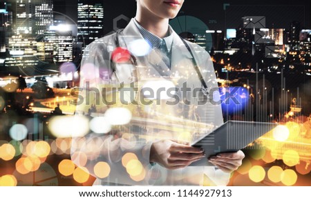 Close up of young medical industry employee standing against night city view and holding tablet in hands. Young female doctor using tablet. Double exposure with media interface icons