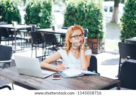 young red-haired woman thinks what to write down in a notebook in a summer cafe. A young woman sits in a cafe and works Royalty-Free Stock Photo #1145058716