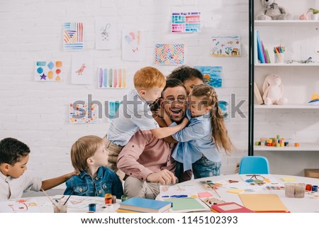 interracial kids hugging happy teacher at table in classroom Royalty-Free Stock Photo #1145102393