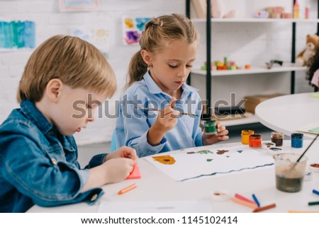 focused preschoolers drawing pictures with paints and paint brushes at table in classroom