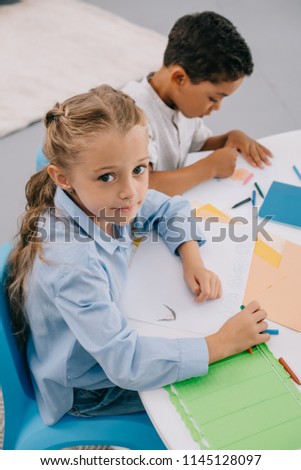high angle view of caucasian kid looking at camera while sitting near african american classmate at table in classroom