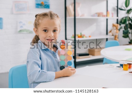 portrait of cute child showing paints in hands while sitting at table in room