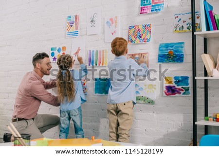 smiling teacher helping little preschoolers hang colorful pictures on wall in classroom