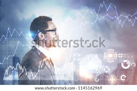 Side view of a young handsome African American businessman wearing glasses and a gray suit. Growing graphs and HUD against a gray city background. Toned image double exposure Royalty-Free Stock Photo #1145162969