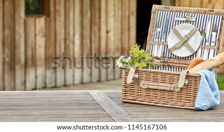 Open vintage fitted picnic hamper with baguettes outdoors on a rustic table near a wooden cabin with space alongside for placement of food in panorama format