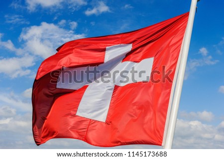 The swiss flag is waving in the wind against a blue sky with clouds