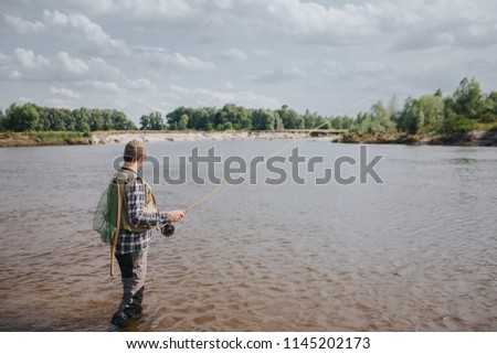 A picture of man standing in water and fishing. He is holding spinning in hands and looking at water. Guy has fishing net on the back. He looks calm and concentrated.