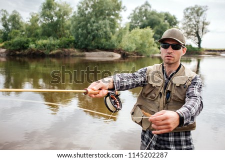 A picture of cool guy stands in water and fishing. He holds fly-fishing with reel under it in one hand and spoon from it in the other one. Guy looks straight. He wears sunglasses.
