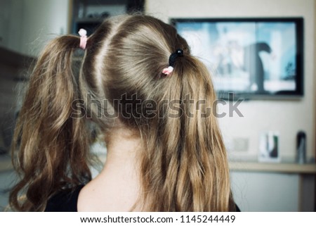 The Little blonde girl with two ponytails on her head is watching cartoons in the kitchen