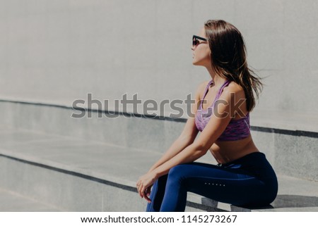 Sideways shot of attractive slim woman in sportsclothes, wears sunglasses, sits on stairs, looks aside, thinks about something, has outdoor training in morning. People, lifestyle and sport concept