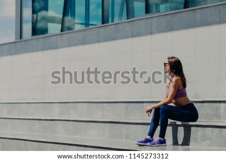 Horizontal shot of beautiful sporty woman in casual clothes, sits on stairs, has rest after gymnastics, poses outdoor. Female athlete in sneakers trains in open air. People, active lifestyle concept