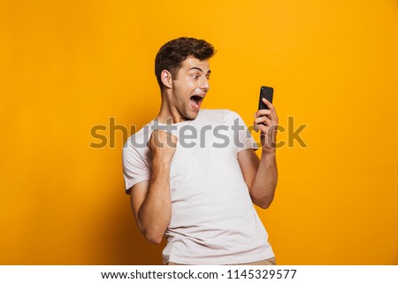 Portrait of an excited young man looking at mobile phone isolated over yellow background, celebrating